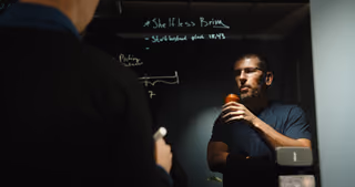 Kollegor diskuterar ett problem på varsin sida av en skrivtavla av glas.
